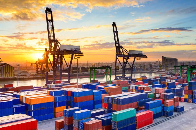 Grandes guindastes e contêineres de carga Exportação coloridos em um porto comercial movimentado durante o pôr do sol.